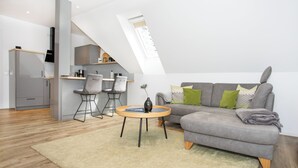 Ein moderner Wohnraum mit einer offenen Küche im Loft-Stil, einem grauen Sofa mit grünen und gemusterten Kissen und einem runden Couchtisch mit einer Vase.