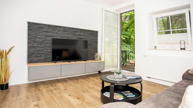 Una sala moderna con un televisor fijado a la pared de piedra, un sofá gris, una mesa de centro redonda y un ventanal corredero que da a un espacio exterior.
