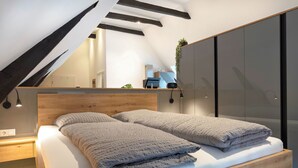 A modern bedroom with a wooden headboard, a large bed with grey bedding, a bedside table, and a built-in wardrobe with a mirror.