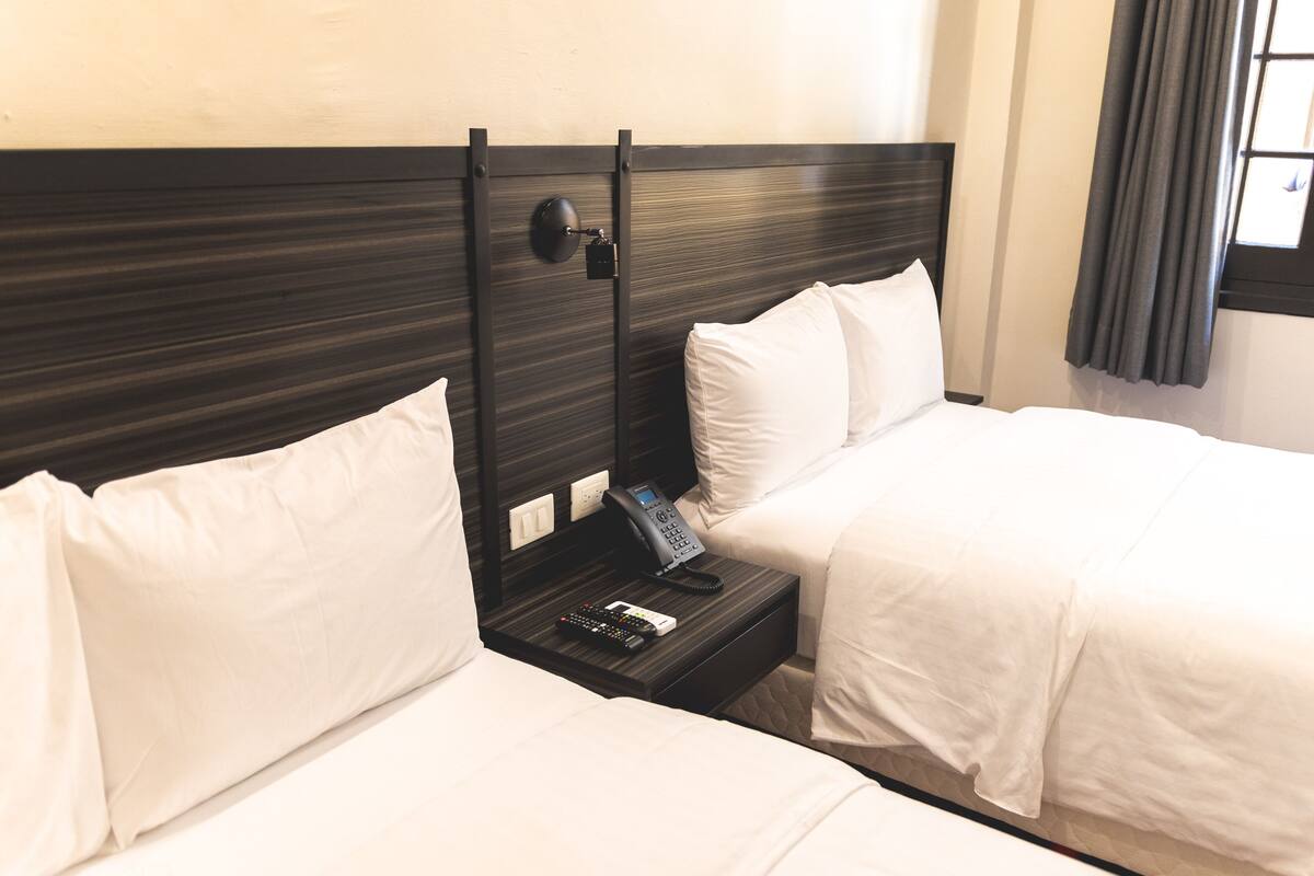 A hotel room with two beds, a nightstand with a phone and remote controls, and a window with curtains.