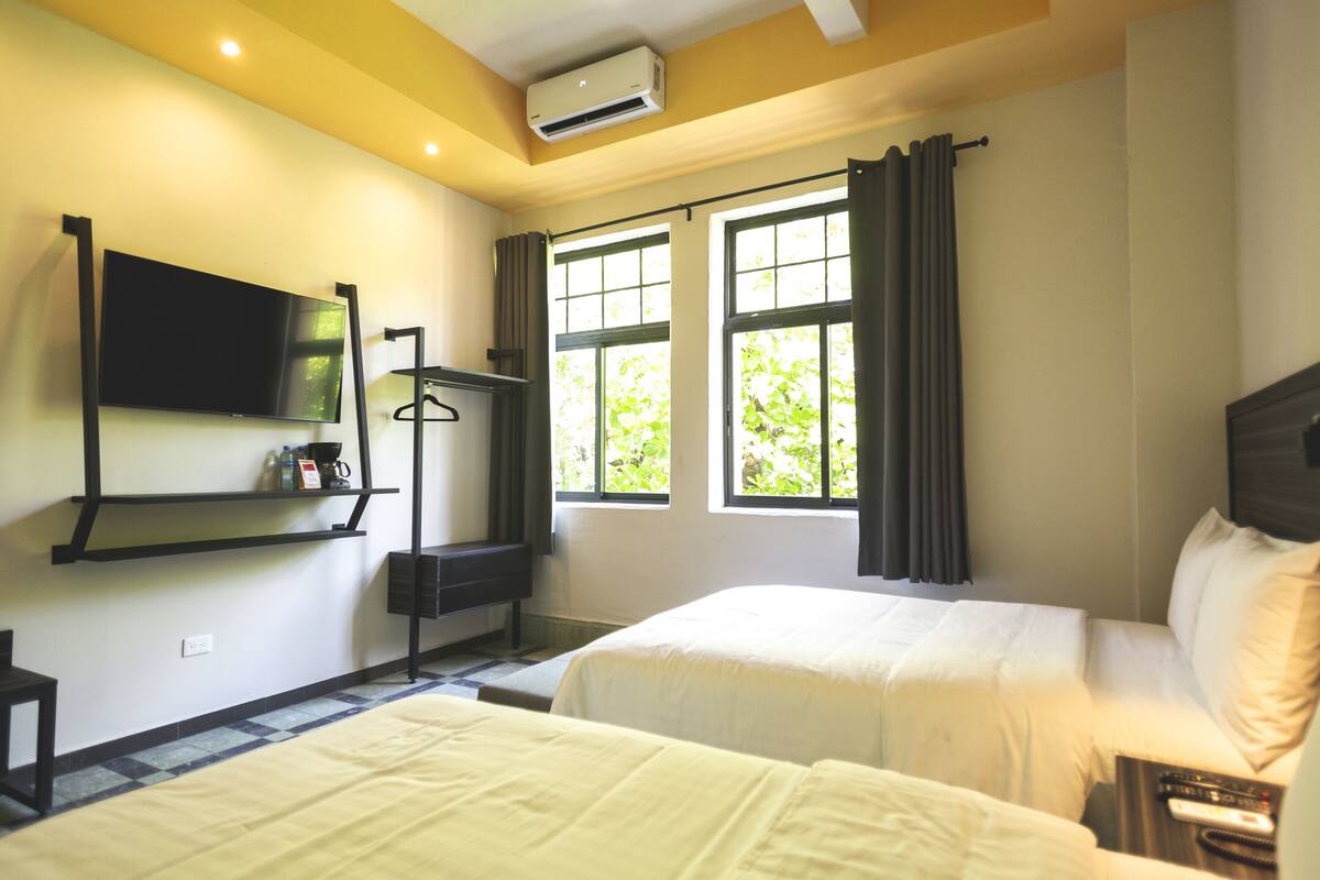 A hotel room with two beds, a TV mounted on the wall, a window with curtains, and a small table with a remote control.