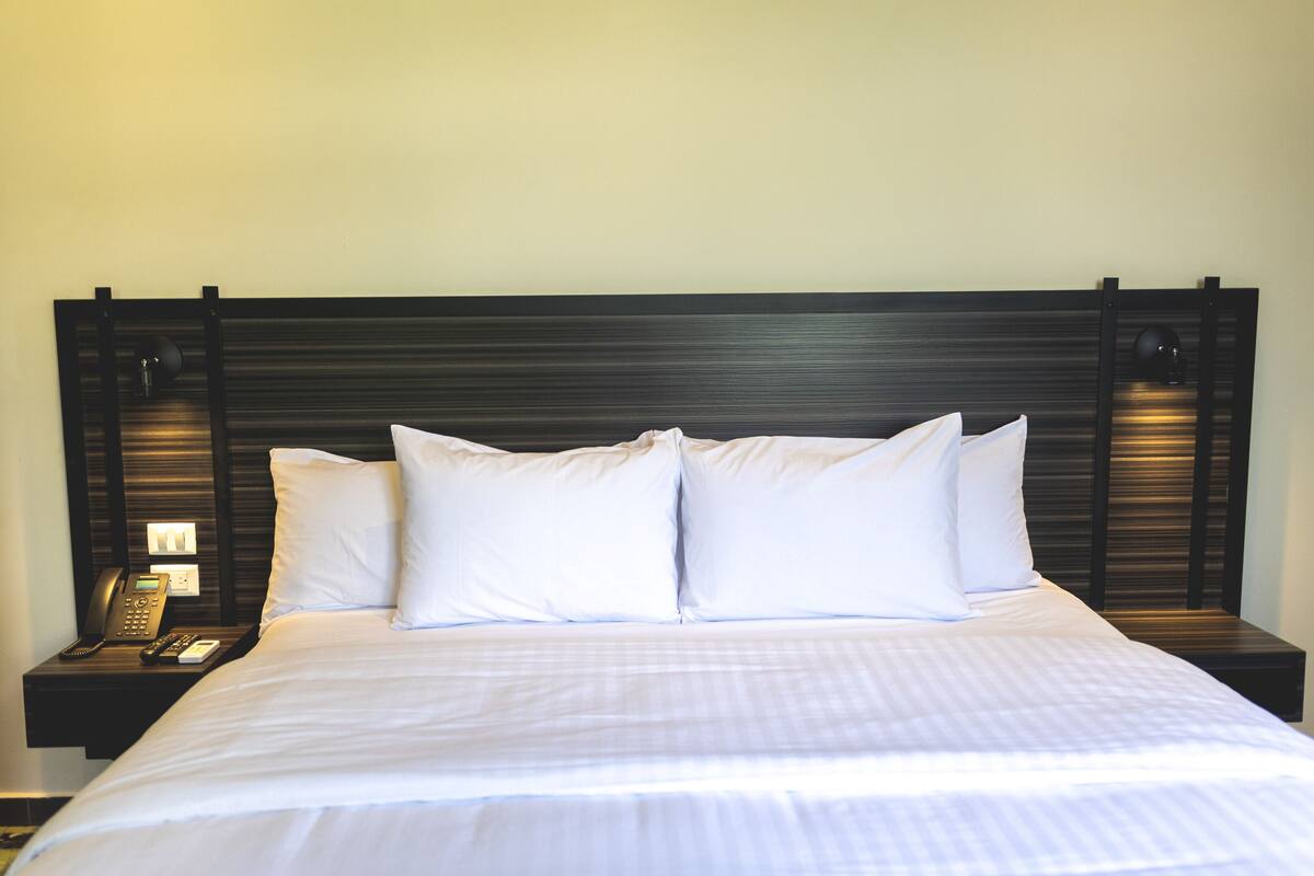 A neatly made bed with white linens and pillows, a bedside telephone, and a nightstand with a lamp.
