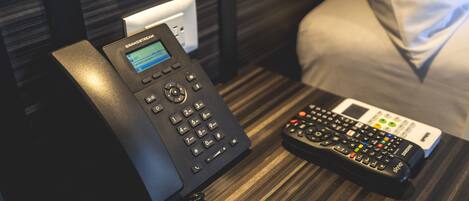 A hotel room with a telephone, a remote control, and a power outlet on the wooden nightstand.