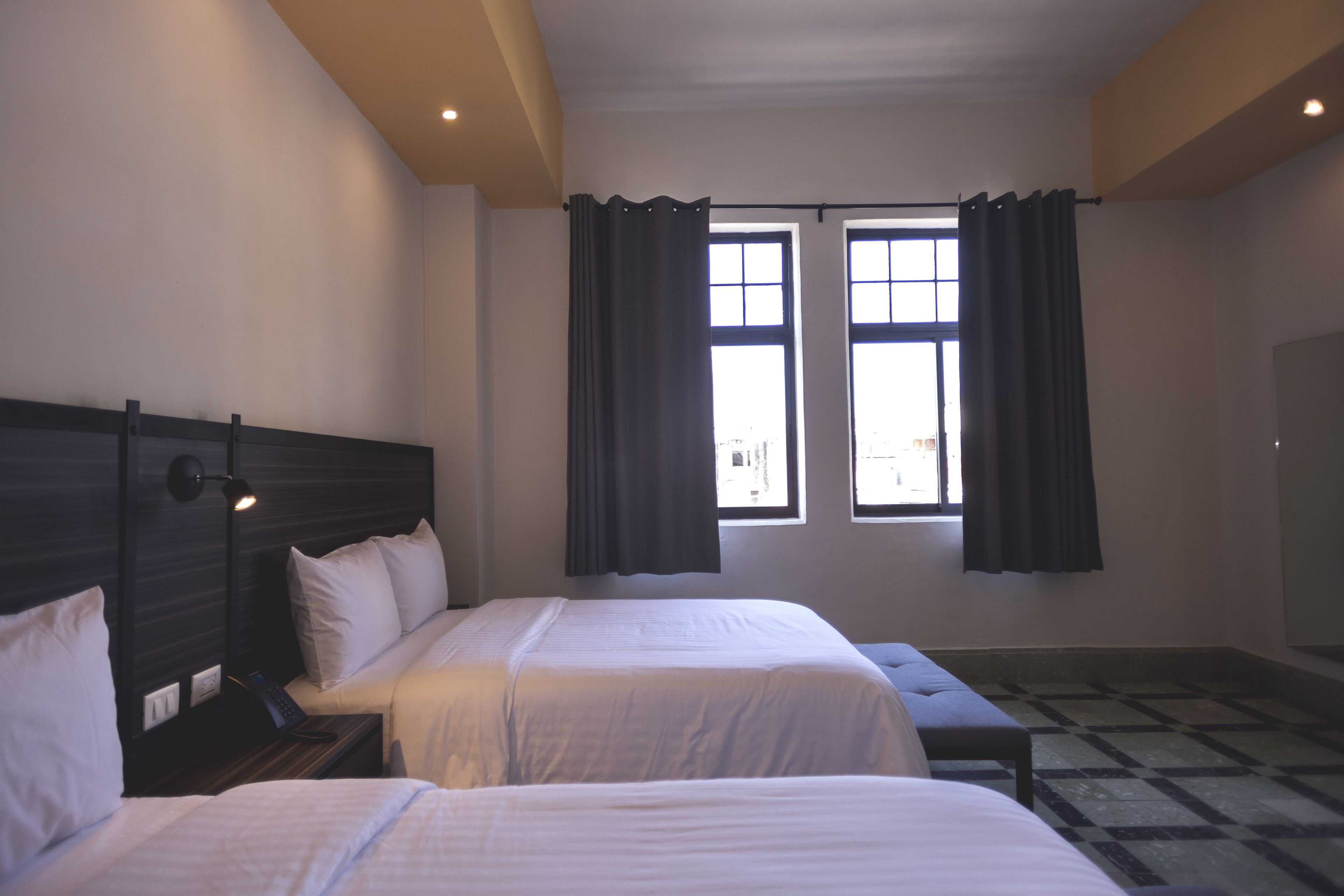 A hotel room with two beds, a nightstand, a lamp, and a large window with curtains.