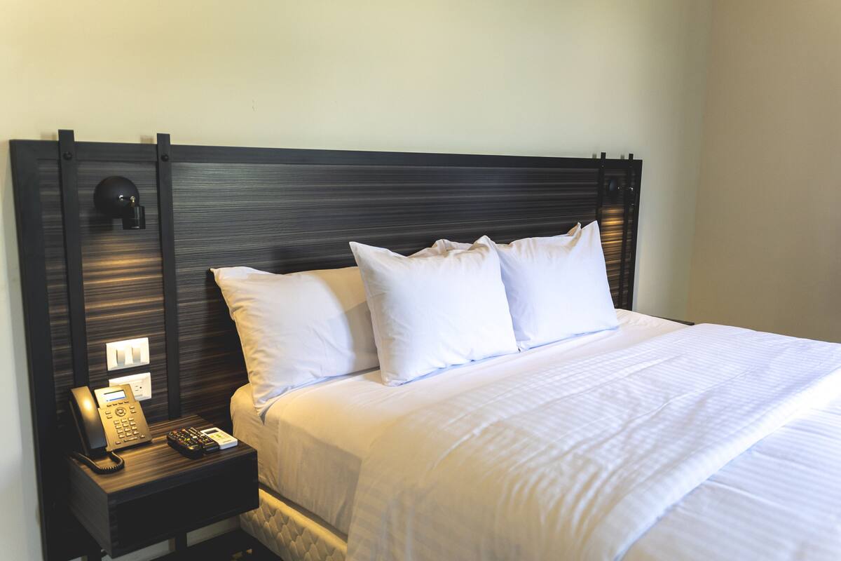 A neatly made bed with white linens and pillows, a bedside telephone, and a wall-mounted light fixture.