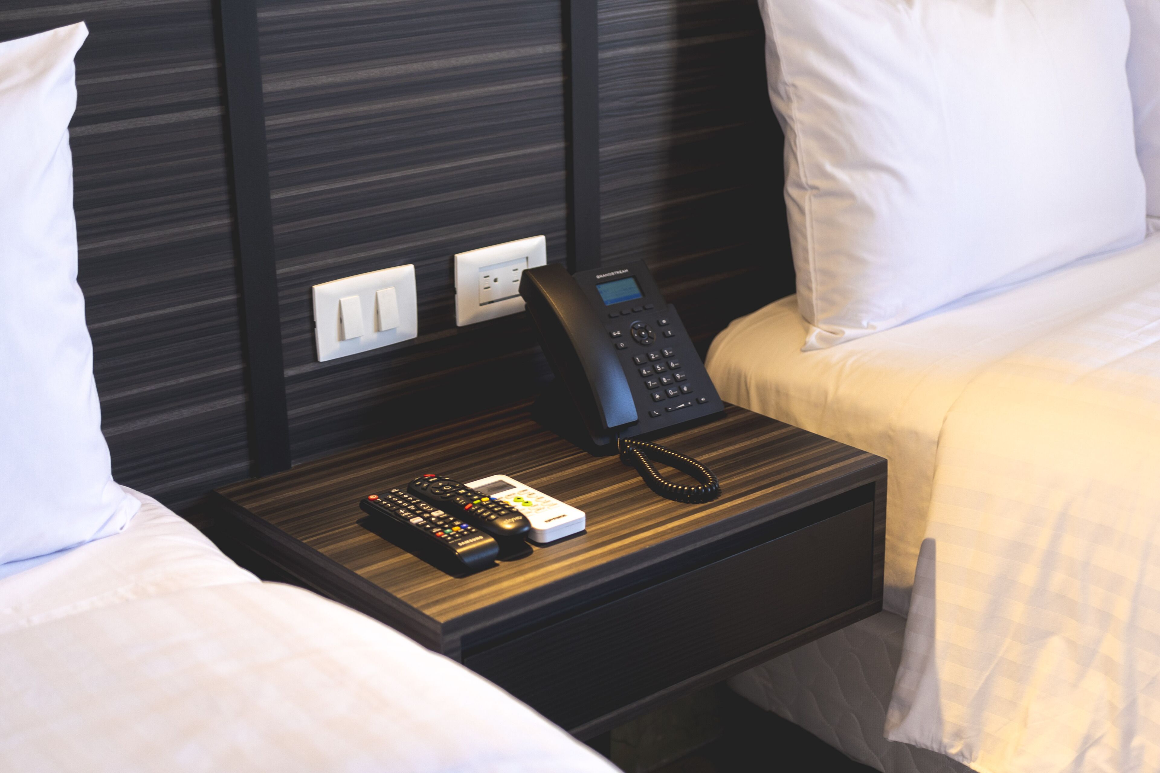 A hotel room with a bedside table featuring a telephone, a remote control, and a power outlet.