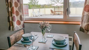 A dining area with a table set for two, a vase with flowers, and a view of the outdoor seating area through a window.