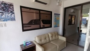 A room with a beige sofa, a small wooden side table with a bottle and a book, a wall-mounted air conditioner, and a poster of a car. - OYO 869 Jnv Dream Hotel (Subic)