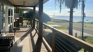 A wooden deck with a railing overlooking a body of water, a green building, and a palm tree. - Wedgewood on the Bay • Waterfront Retreat (Seadrift)