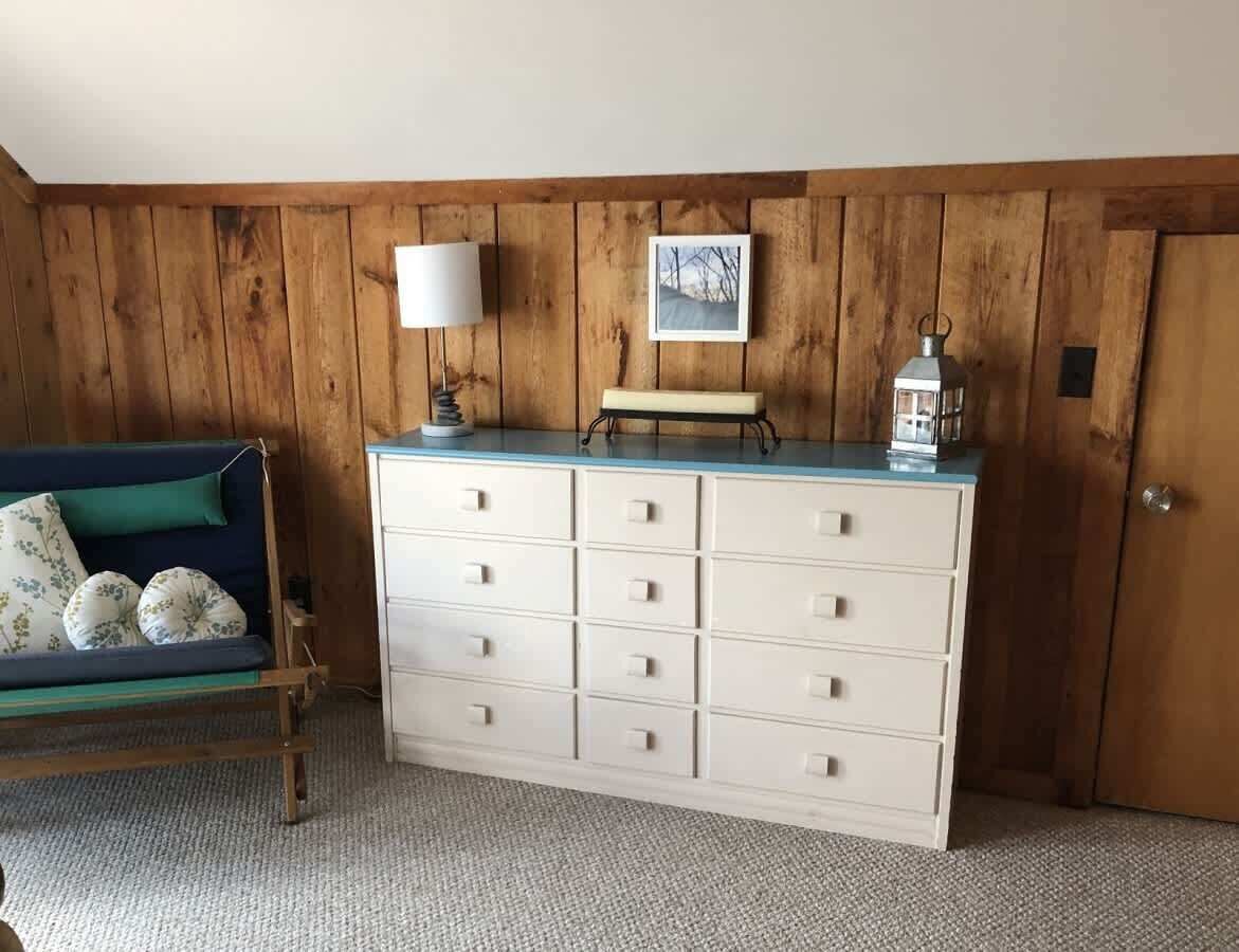 Une pièce avec un mur lambrissé en bois, une commode blanche dotée de nombreux tiroirs, un canapé bleu agrémenté de coussins à motifs floraux, une lampe blanche et un tableau encadré.