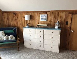 A room with a wooden paneled wall, a white dresser with multiple drawers, a blue sofa with floral cushions, a white lamp, and a framed picture.