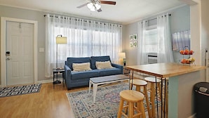 A living room with a blue sofa, a wooden coffee table, a ceiling fan, a dining area with stools, and a kitchen area with a fruit basket.