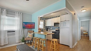A modern kitchen with white cabinets, a microwave, and a refrigerator. There is a dining area with a bench and stools, and a colorful rug on the floor.