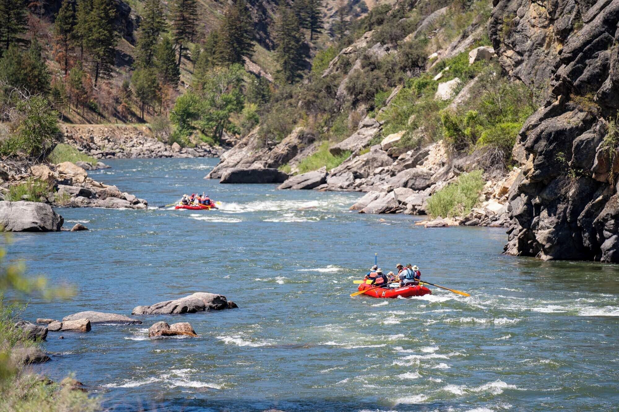 A river with two red inflatable rafts navigating through it, surrounded by rocky terrain and dense greenery.