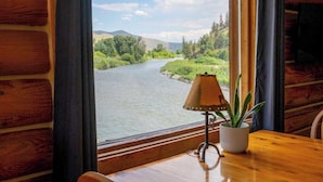 A wooden cabin interior with a large window overlooking a river and forest. A table with a lamp and a potted plant is visible. - River's Fork Lodge (North Fork)
