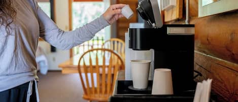 A person pouring coffee into a cup from a coffee machine in a room with wooden furniture and a window.