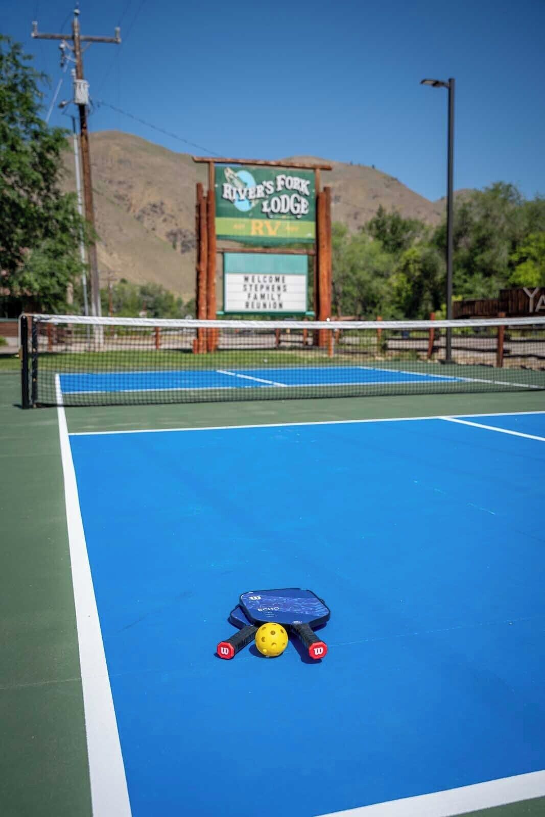 Una cancha de tenis con red, rodeada por un letrero de River's Fork Lodge.