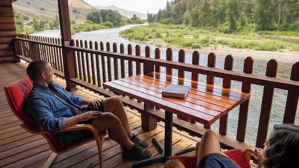A wooden deck with two rocking chairs and a table, overlooking a river and forested hills. - River's Fork Lodge (North Fork)