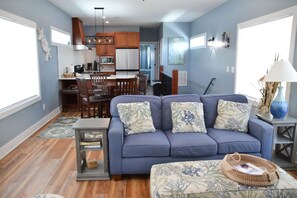 Relax in this inviting living room with a sofa, side table, and woven basket, surrounded by a large window and a kitchen area.
