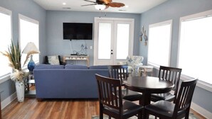 A living room with a blue sofa, wooden dining table, and chairs, a ceiling fan, and a TV mounted on the wall. - Unique 3BR Home with great views Beach Access (Atlantic Beach)