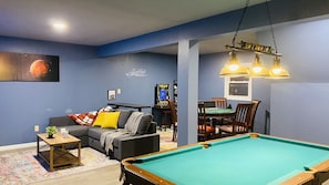 A room with a pool table, a sofa with cushions, a small table with a plant, and a game console.