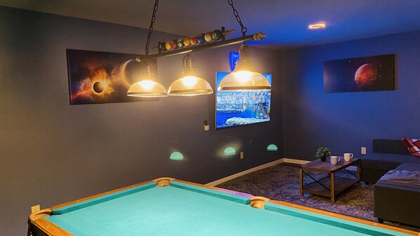 A pool table with overhead lighting, a television displaying a cityscape, and a billiards table with a pool cue. - NEW!! Pocono!! Family Friendly, Hot tub, Fireplace (Tobyhanna)