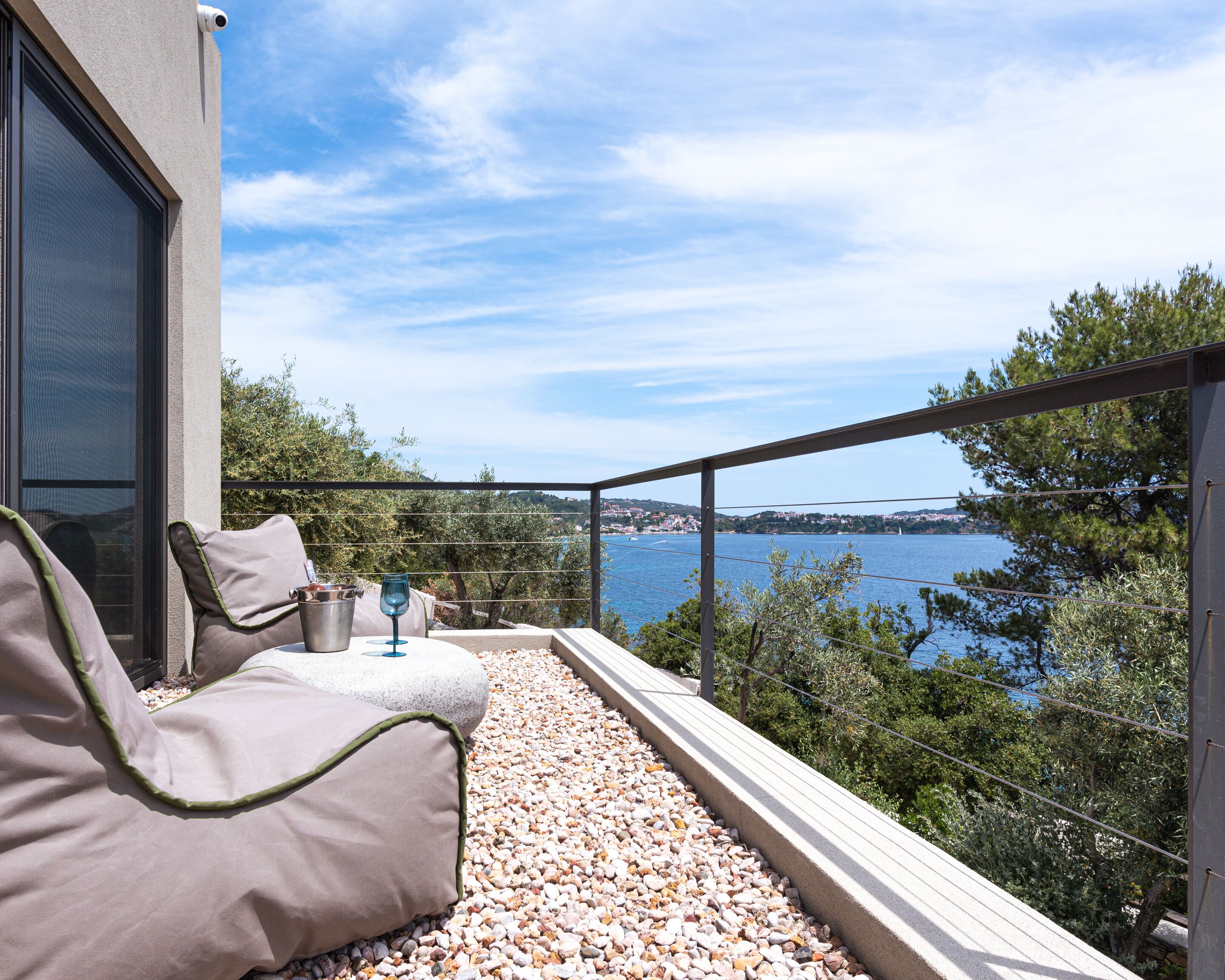 A balcony with a sofa, a glass, and a stone table, overlooking a body of water and a distant town.