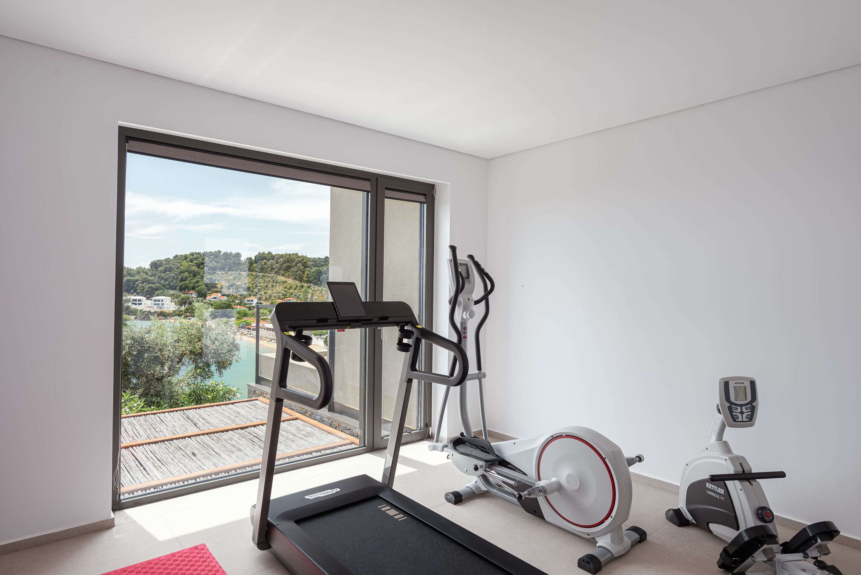 A modern room with a treadmill, elliptical, and stationary bike, large windows with a view of a deck and greenery.