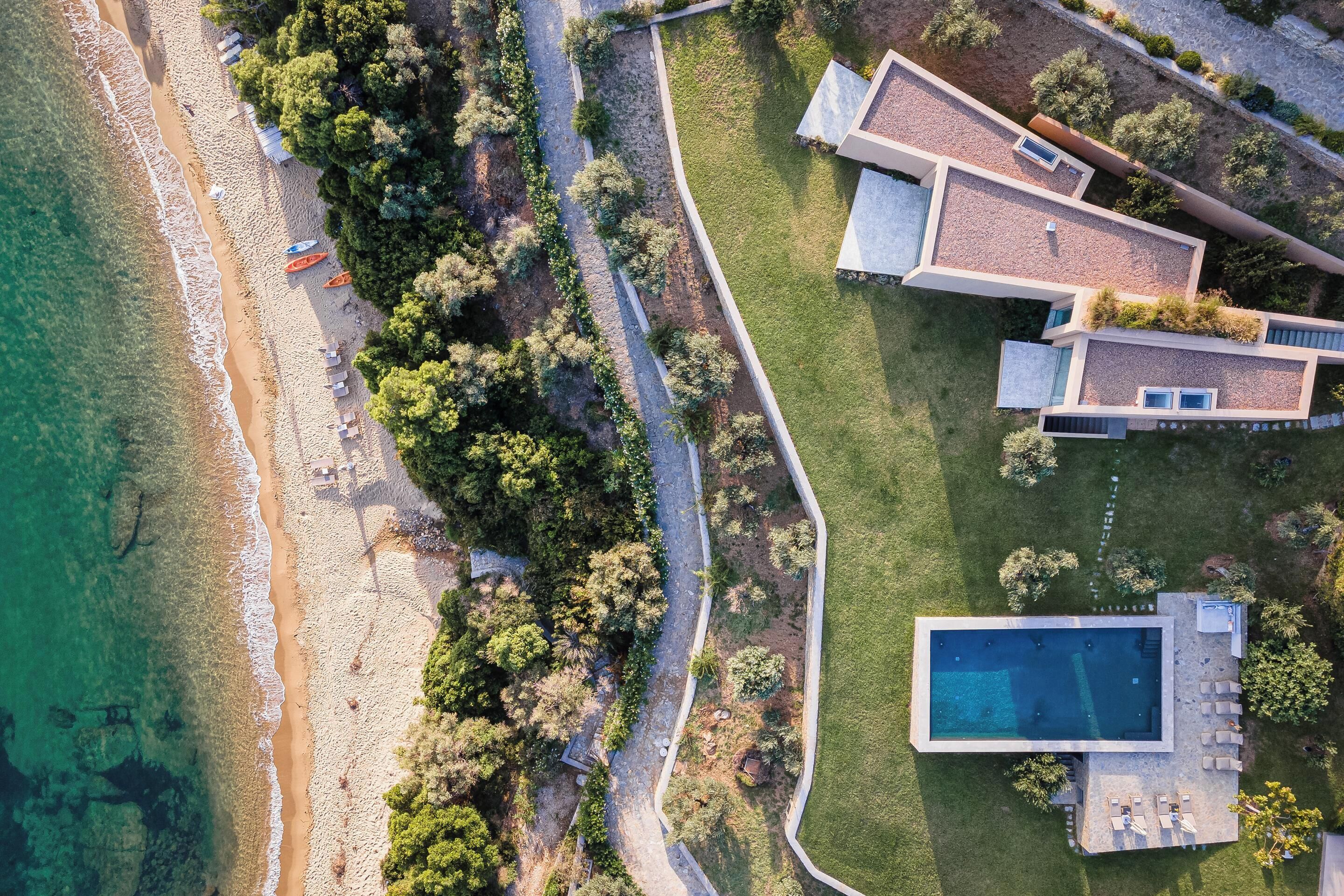 Ett flygbildsfoto av en fastighet vid kusten med en swimmingpool, en strand och moderna hus.