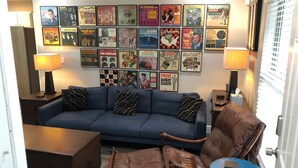 A living room with a blue sofa, brown leather armchair, and a small round table. A wall of framed album covers is visible. - Amazing Five-Star Luxury Thornton Park two Bedroom Apt - close to Kia Center. (Orlando)