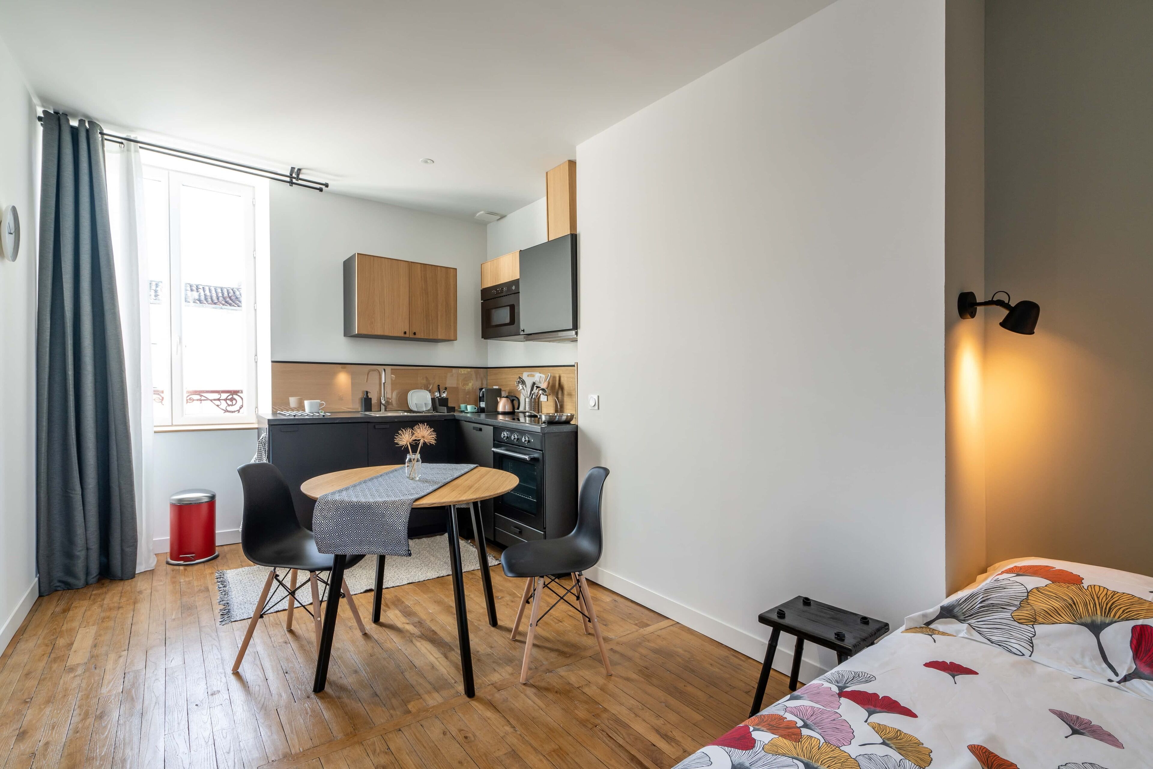 Een ruimtelijk ingerichte leefruimte met een kitchenette, een eethoek met een tafel en stoelen, en een bed met een bloemenprint deken.