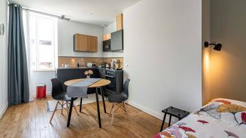 Een ruimtelijk ingerichte leefruimte met een kitchenette, een eethoek met een tafel en stoelen, en een bed met een bloemenprint deken.