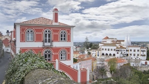 A red and white building with arched windows and a balcony, surrounded by other buildings and a cloudy sky. - Villa Bela Vista (Sintra)