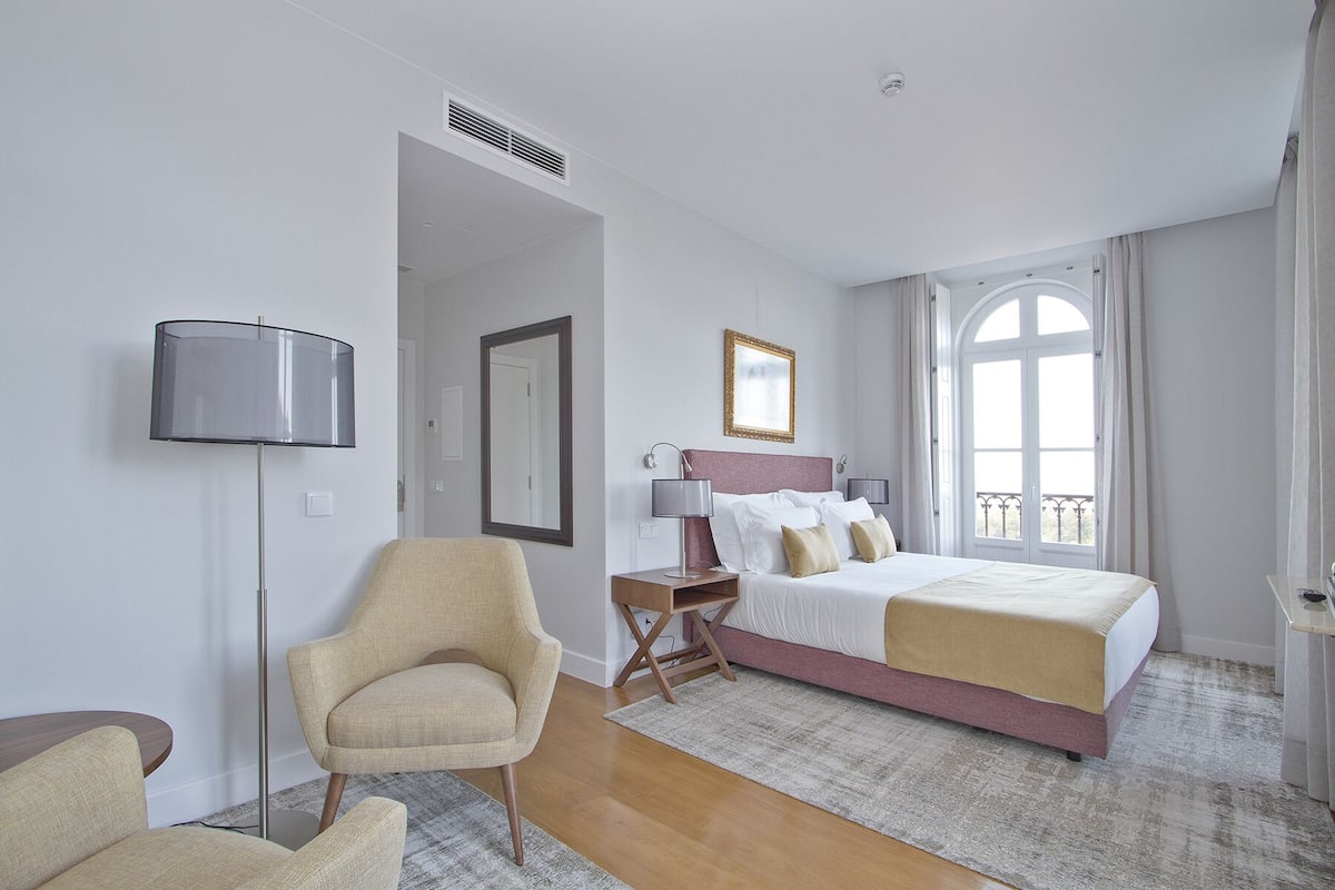 A hotel room with a bed, a chair, a desk, a lamp, a mirror, and a window.