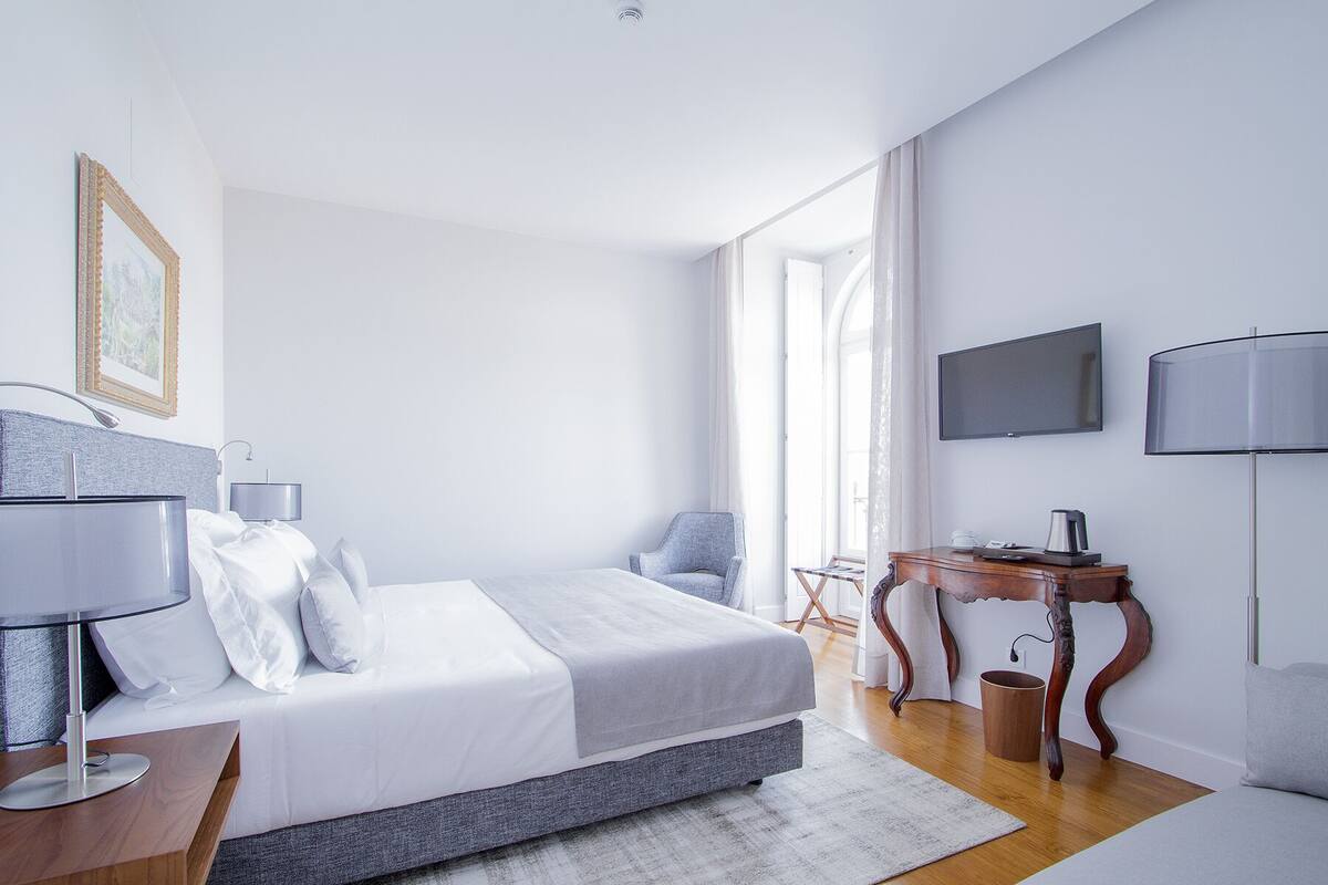 A modern hotel room with a large bed, a desk, a chair, a television, and a window with curtains.