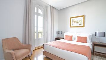 Quarto de hotel com uma cama grande, duas mesas de cabeceira, uma cadeira, janela com cortinas e um quadro na parede.