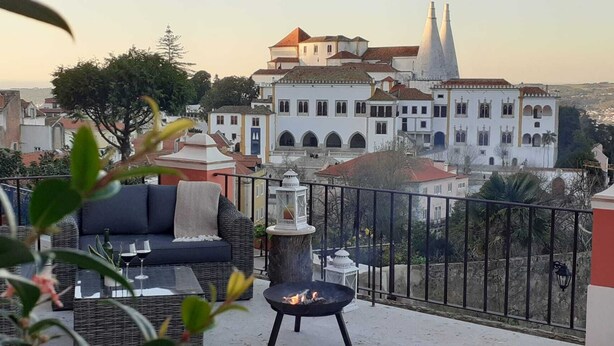 A balcony with a sofa, a small table, and a fire pit. In the background, there are buildings with a church and a tower. - Villa Bela Vista (Sintra)