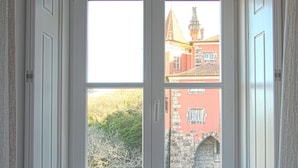 A room with a bay window overlooking a building with a red facade and a tower. There is a bench with cushions in front of the window. - Villa Bela Vista (Sintra)