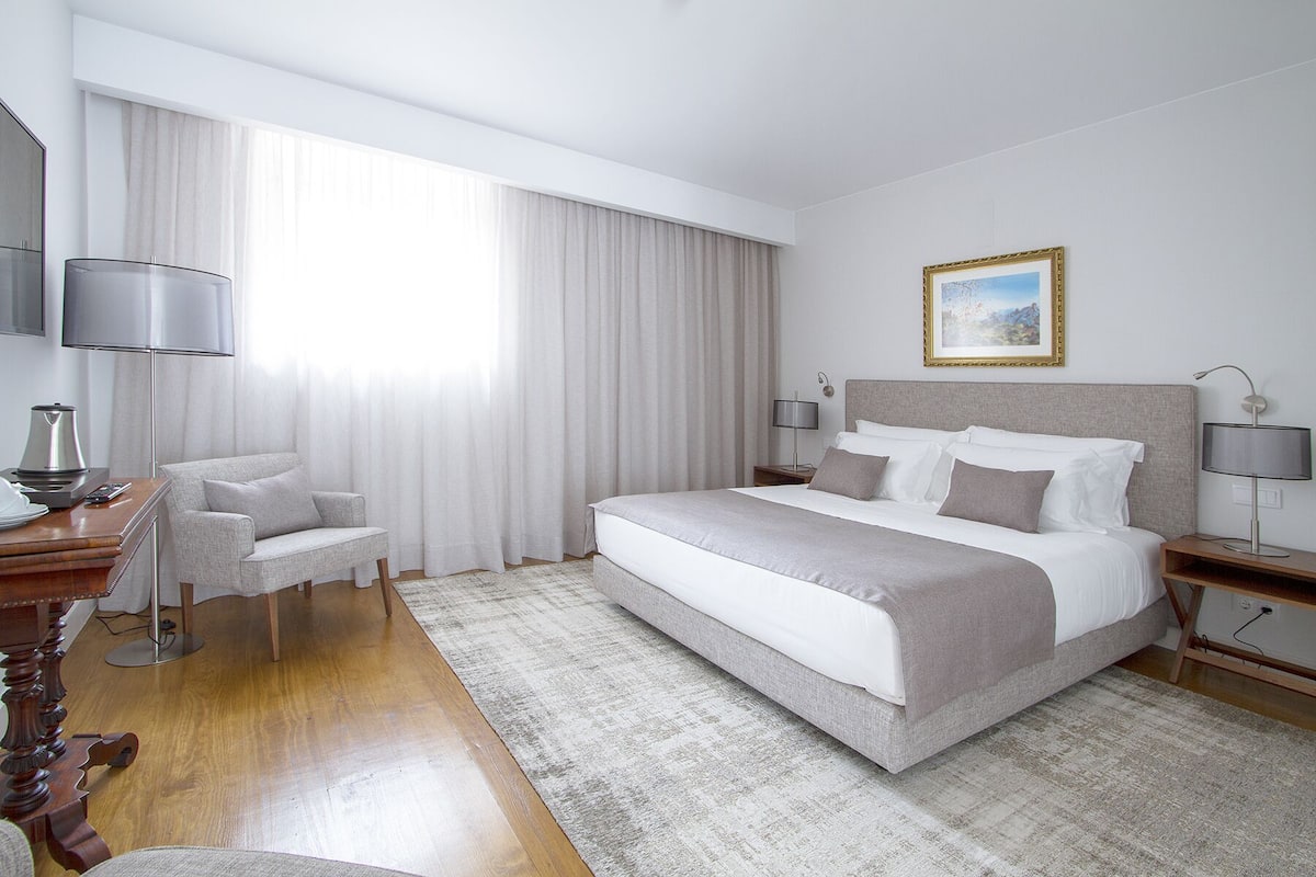 A hotel room with a large bed, a desk with a lamp, a chair, and a painting on the wall.