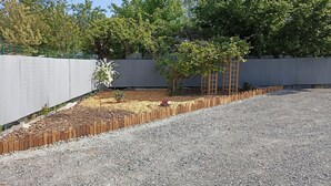 Un chemin de gravier mène à un jardin clos, agrémenté d’arbres et d’une tonnelle en bois.