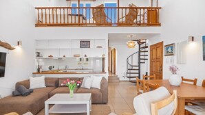 Amplia sala de estar con sofá, mesa de centro y mesa de comedor, una cocina al fondo y una escalera que conduce a un nivel superior.