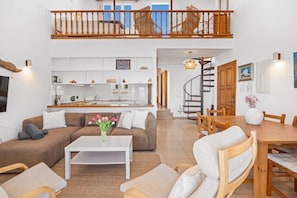 En rummelig opholdsstue med sofa, sofabord og spisebord, et køkken i baggrunden, og en trappe, der fører op til en etage.