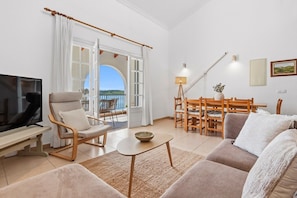A living room with a TV, a sofa, a dining table with chairs, and a balcony with a view.