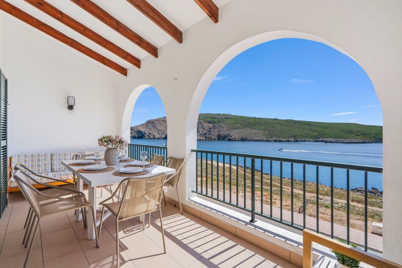 Un balcón con mesa lista para comer, con vista a una masa de agua y un paisaje montañoso.