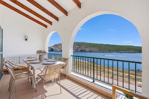 A balcony with a table set for dining, overlooking a body of water and a hilly landscape. - Tramontana 115-P1 - Caressing the Mediterranean (Fornells)