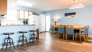 Una cocina moderna con armarios blancos, un comedor con sillas de madera y una mesa, y una zona con taburetes de bar.