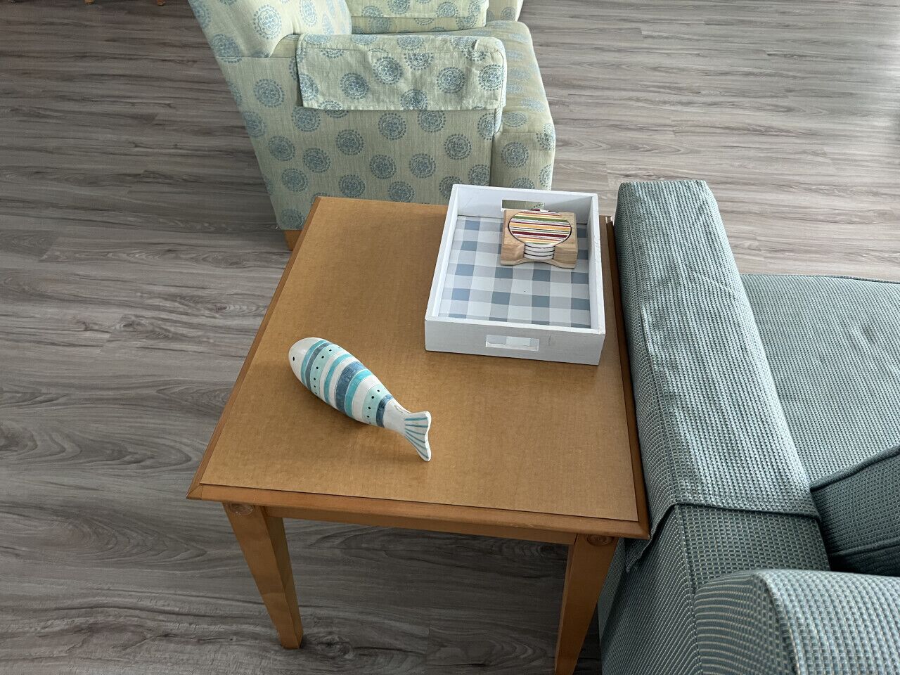 Une petite table en bois, sur laquelle se trouvent une boîte à motifs à carreaux et un jouet en forme de poisson rayé, est placée à côté d’un fauteuil à motifs et d’un canapé recouvert d’un tissu texturé.