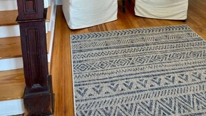 Un escalier avec des marches en bois et des balustrades blanches, deux fauteuils de couleur crème ornés de coussins à motifs, une petite table avec une lampe et un tapis à motifs sur le sol.