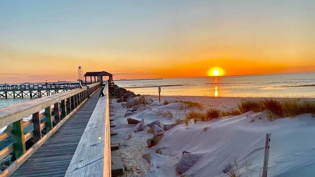 Une promenade en bois menant à une plage bordée de dunes de sable, avec un coucher de soleil en arrière-plan.