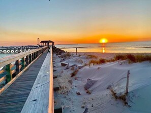 A wooden boardwalk leading to a beach with sand dunes and a sunset in the background.